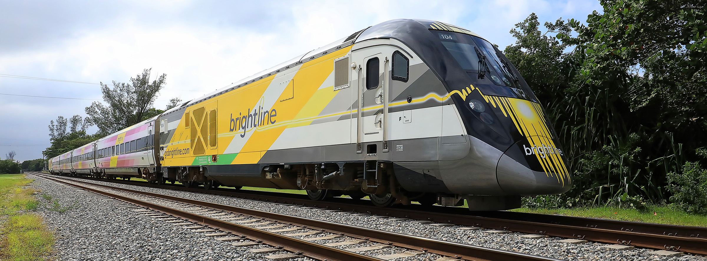 blright line train florida - A sleek, modern Brightline passenger train with a yellow and gray design travels on railroad tracks through a green, tree-lined area under a partly cloudy sky.