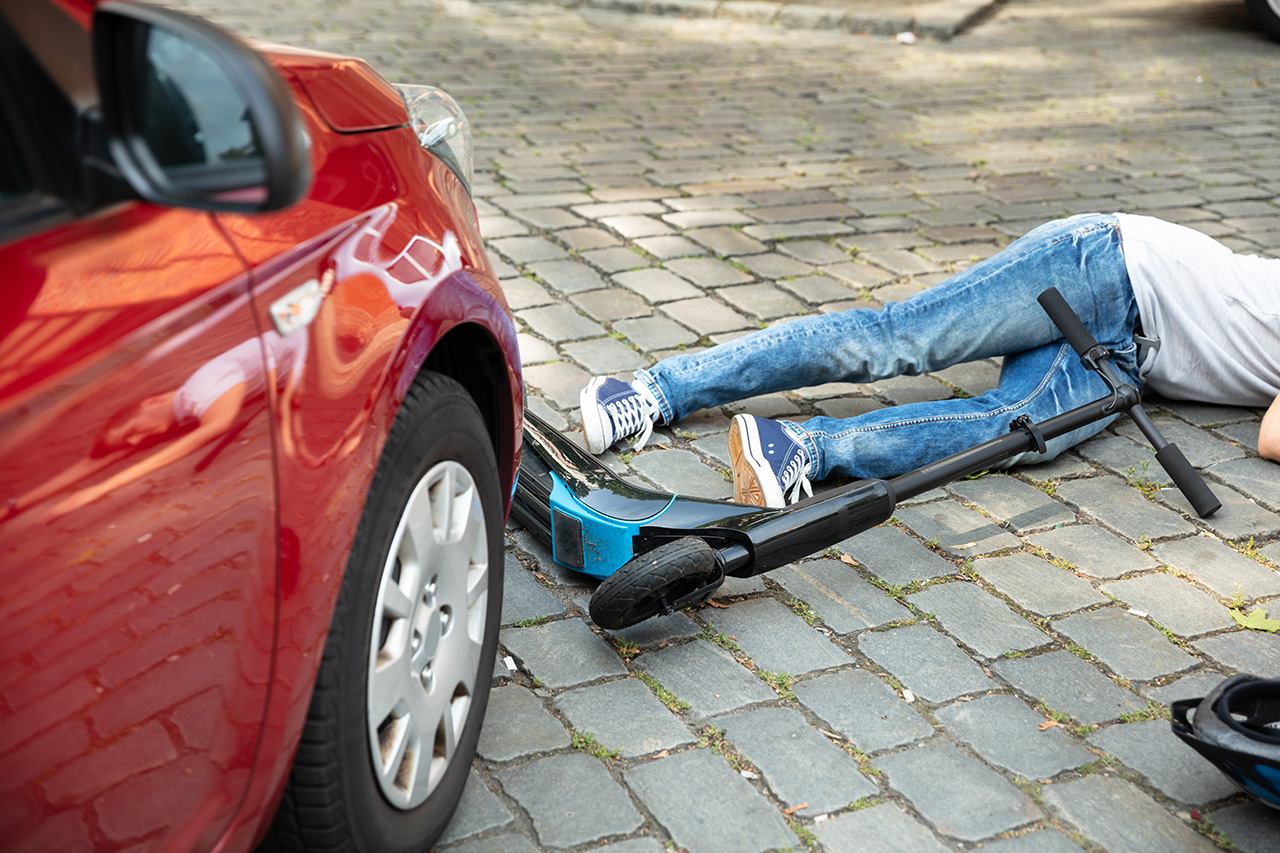 A person lying on cobblestone pavement next to an overturned electric scooter and a red car, suggesting a collision or accident involving both the scooter and the car. Only part of the person’s body is visible.