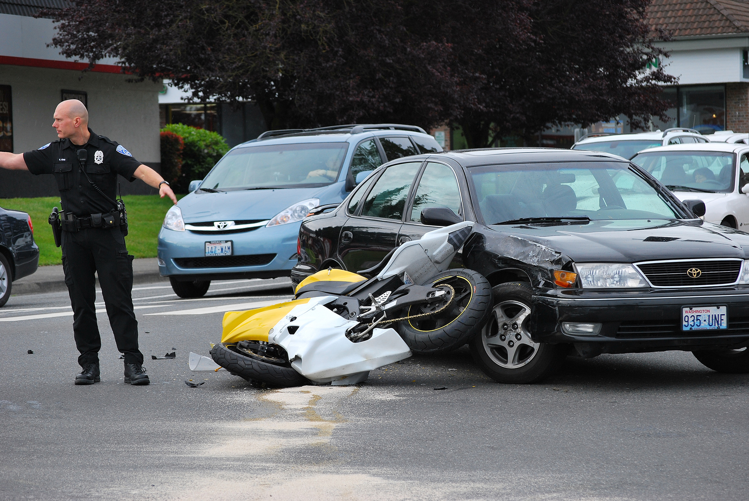 A police officer directs traffic at the scene of a collision between a black car and a yellow-and-white motorcycle lying on its side in the intersection, with other vehicles visible in the background.