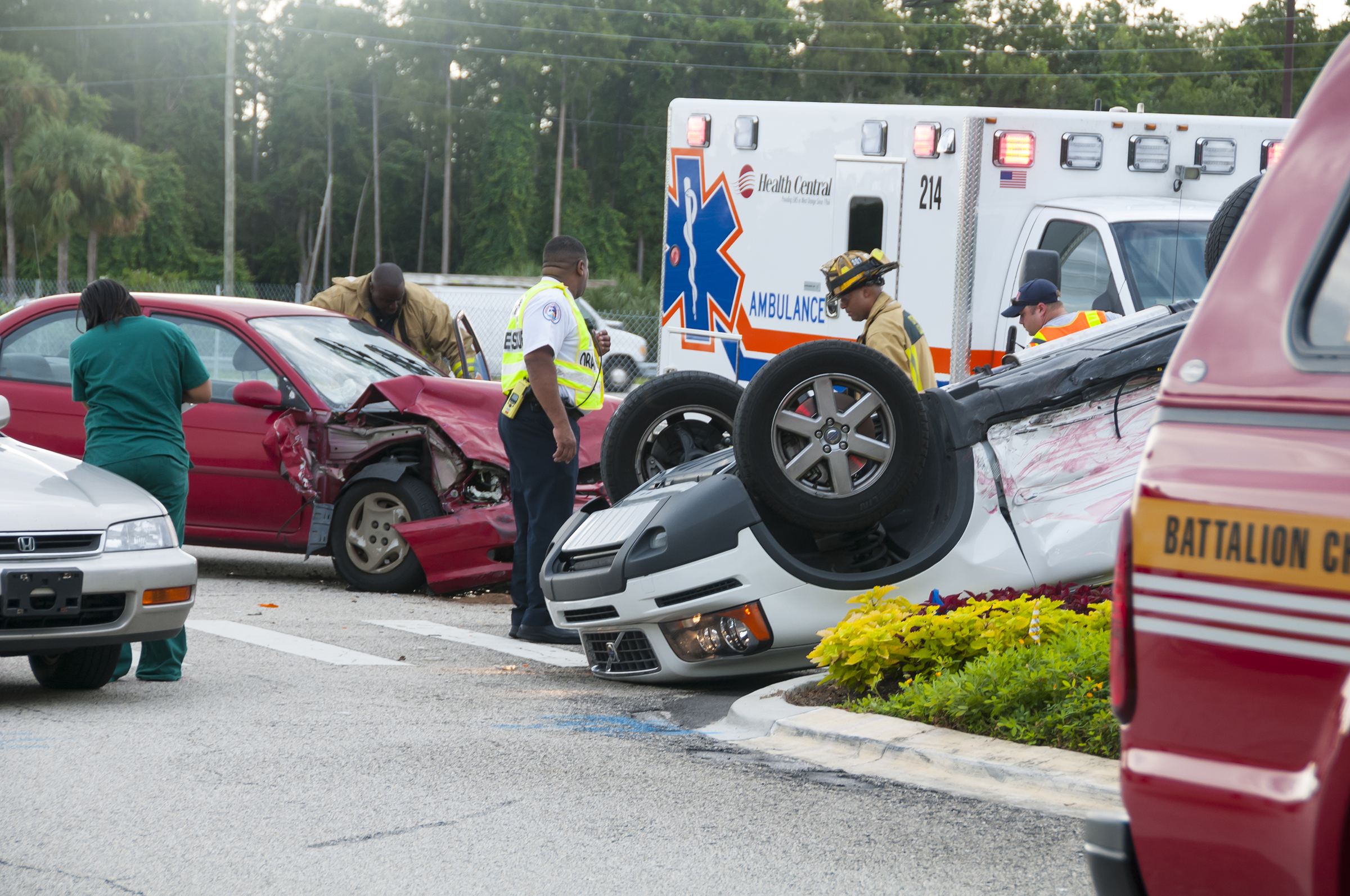  A red car with a damaged front and a white car flipped upside down after a crash; emergency personnel stand nearby at the scene on a road.