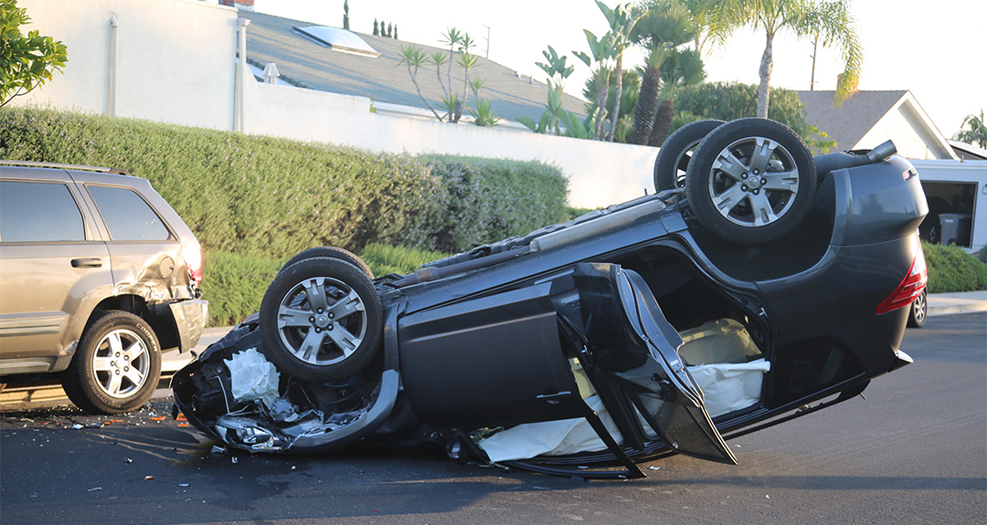 A black SUV is flipped upside down on a suburban street after an accident. The car’s airbags have deployed, and another parked SUV nearby shows damage to its rear. Palm trees and houses are visible in the background.