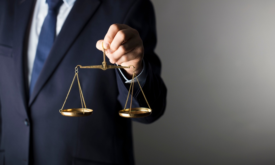 Show thumbnail preview A person in a dark suit and blue tie holds a brass balance scale in one hand, symbolizing justice and fairness, against a plain light grey background.