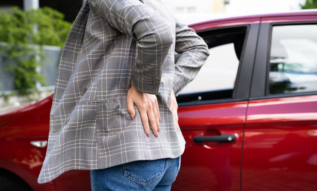A person in a plaid jacket stands beside a red car, holding their lower back with one hand, appearing to be in pain or discomfort.