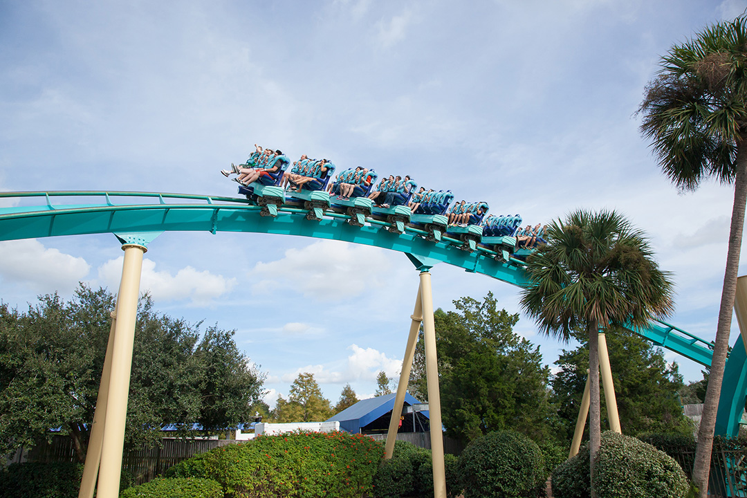 A teal roller coaster train filled with passengers speeds along a raised track, surrounded by green trees and bushes under a partly cloudy sky.