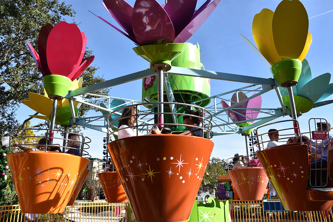 Colorful flower-shaped ride at an amusement park with people sitting inside petal-themed cars. The ride is outdoors under a blue sky with trees visible in the background.