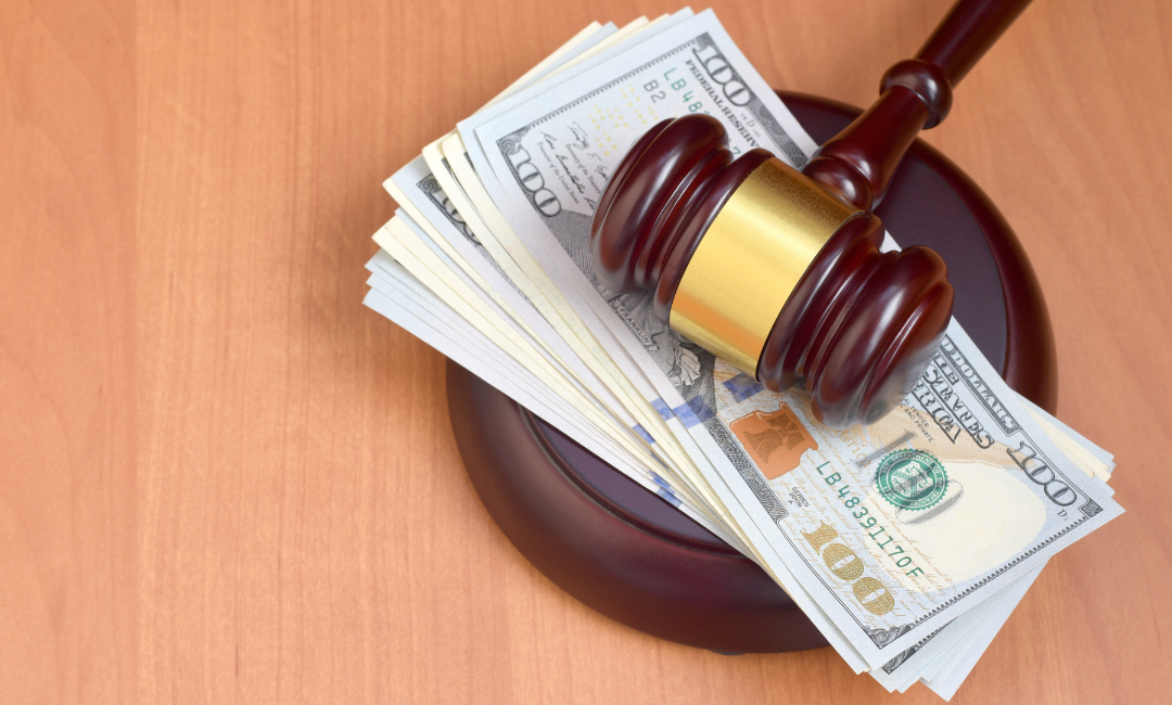 A judge’s gavel rests on top of a stack of hundred-dollar bills on a wooden surface, symbolizing legal decisions involving money, fines, or financial settlements.
