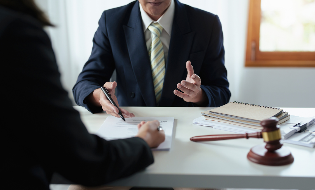 Two people in business attire sit at a desk discussing documents. One gestures while the other writes. A gavel, notepad, and files are on the table, suggesting a legal or business meeting.