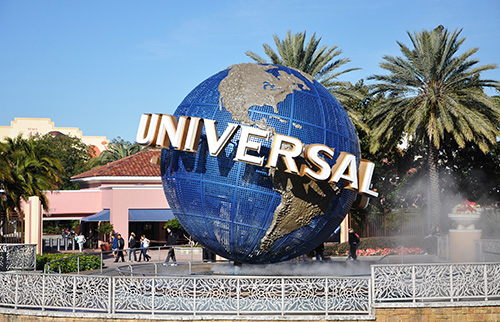 A large blue globe sculpture with the word UNIVERSAL in bold white letters stands at the entrance of Universal Studios, surrounded by palm trees, water features, and people walking by.