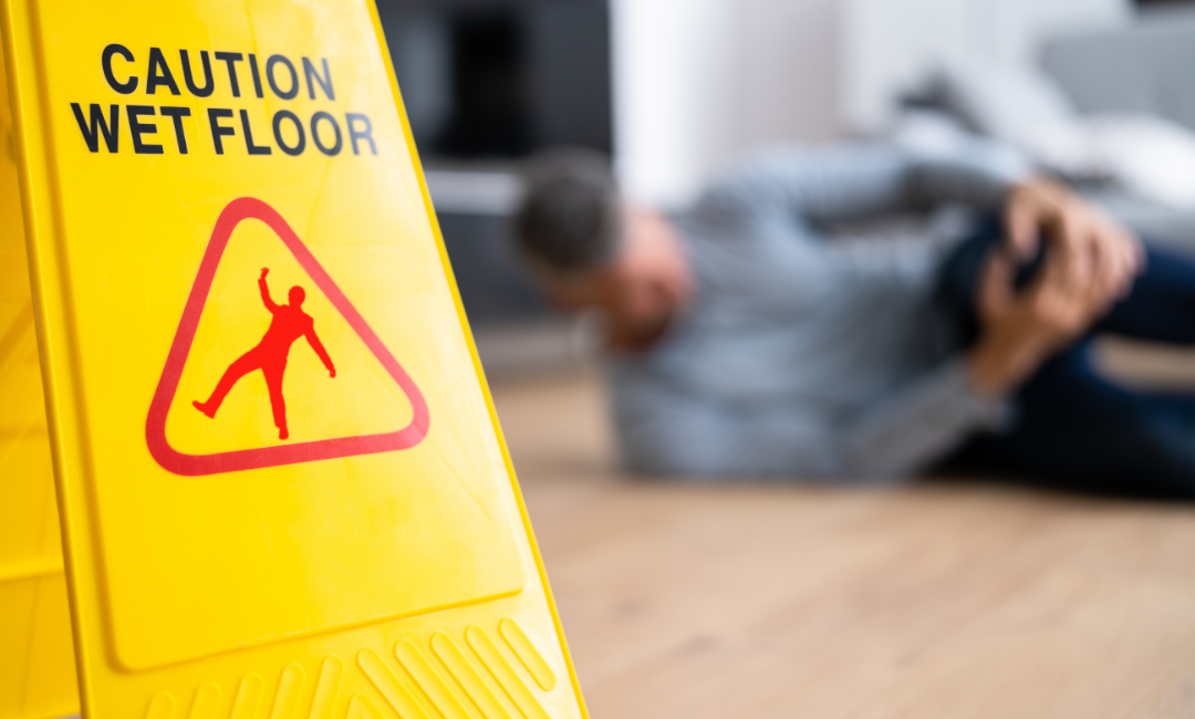 A yellow caution sign reading Caution Wet Floor is in focus, while a person in the background sits on the floor, holding their knee as if injured from a fall.