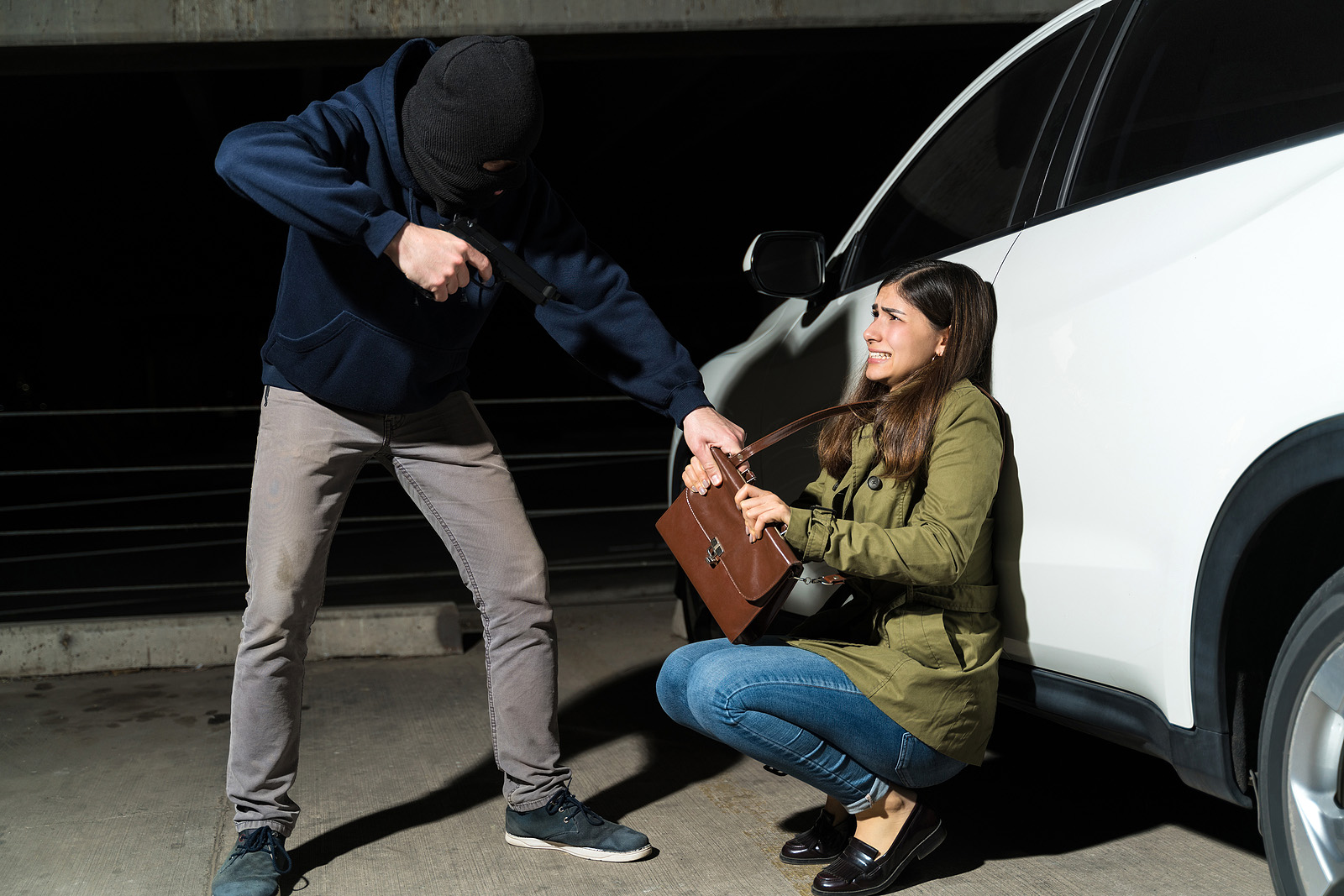  A person wearing a mask and pointing a gun threatens a woman crouching by a white car, grabbing her purse while she looks frightened—an example of negligent security.