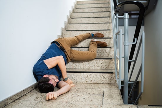 An unconcious man is lying on his side at the bottom of a staircase, appearing to have fallen. He is wearing a blue shirt, tan pants, and brown shoes, with one arm outstretched and his head resting on his arm.