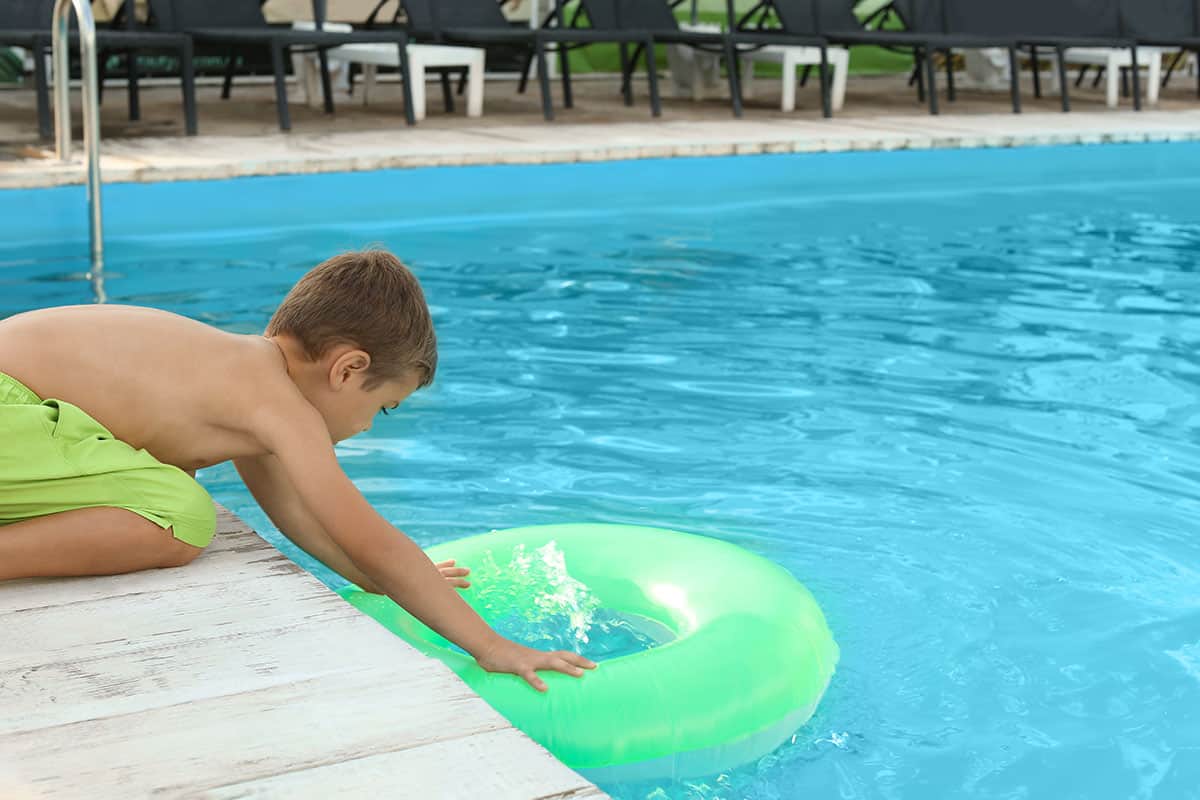 Little child on swimming pool deck reaching for inflatable tube in the pool