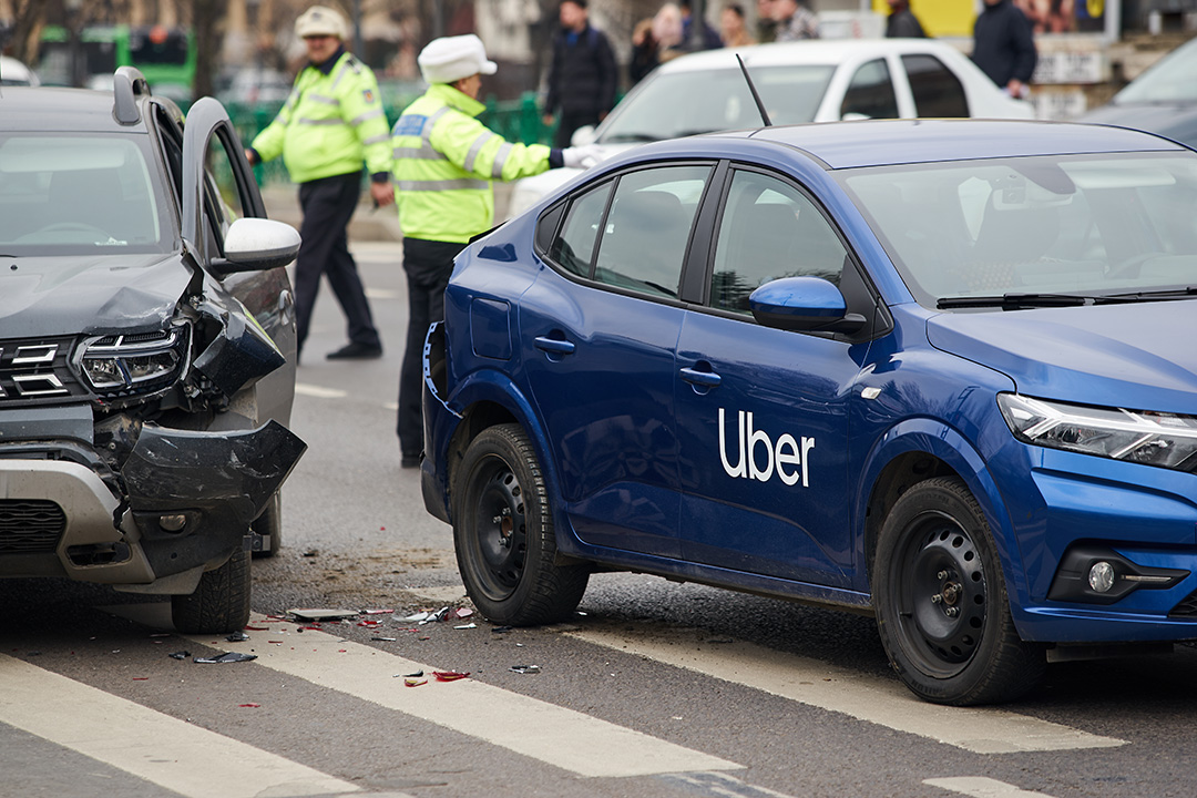 Show thumbnail preview A damaged blue Uber car and a black SUV after a collision at an intersection, with police officers in reflective jackets investigating the accident in the background.